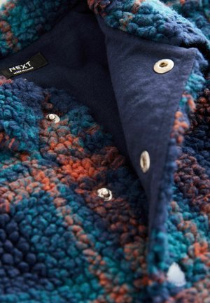 Blau- und orangefarbene strukturierte Fleecejacke mit einem geknöpften Kragen, die eine weiche, flauschige Außenseite und eine dunkelblaue Innenschicht aufweist. Markenlabel sichtbar.