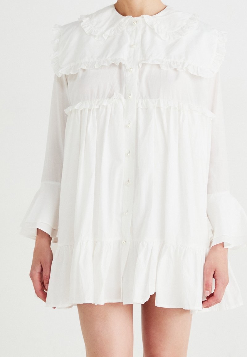 Robe longue blanche à manches longues et fluide, avec grand col à volants, boutons sur le devant et ourlet à volants, portée par une personne les mains détendues le long du corps.