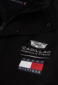 Μαύρο μπουφάν με λογότυπο της ομάδας Cadillac Formula 1 πάνω από το λογότυπο της σημαίας Tommy Hilfiger, με λεπτομέρειες φερμουάρ και κουμπιού.