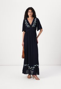 Navy maxi dress with short flutter sleeves, V-neck, and floral embroidery. Smooth texture and flowy design, paired with a brown handbag.
