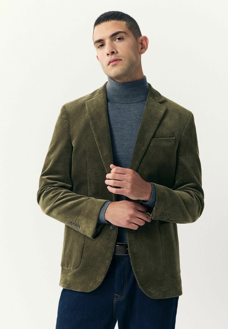 Hombre ajustando un blazer de pana color verde oliva sobre un suéter de cuello alto gris y jeans azul oscuro frente a un fondo blanco liso.