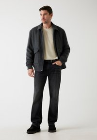 Jachetă bomber din lână gri, cu două buzunare frontale, purtată peste o cămașă crem, asociată cu blugi negri și adidași negri. Design și croială simple.