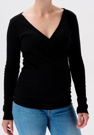 Kobieta w czarnej bluzce z długim rękawem i dekoltem w serek oraz jasnoniebieskich dżinsach, stojąca na białym tle.