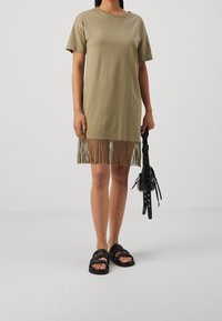 Kvinna klädd i en kortärmad olivgrön klänning med fransar i kanten, håller en svart handväska och bär svarta spännbandsandaler.