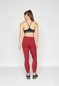 Črn športni modrček z belim logotipom, v kombinaciji z visokovzdignjenimi maroon tajicami. Model nosi črne športne čevlje. Gladek, oprijet material.