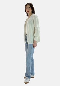 Witte gebreide cardigan met een ontspannen pasvorm, gestructureerde stof en geribbelde boorden, gedragen over een licht topje en gecombineerd met lichtblauwe spijkerbroek en sneakers.