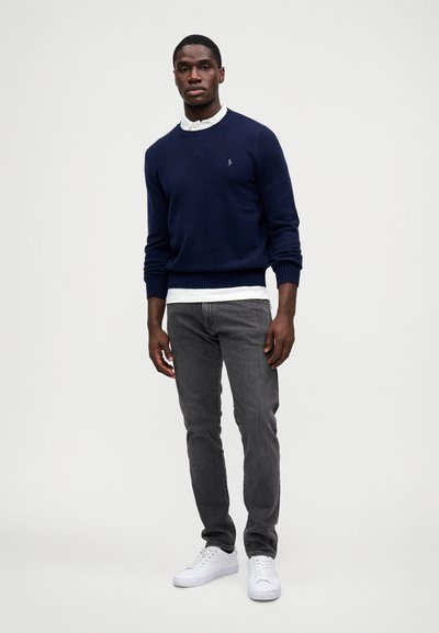 Sweater azul-marinho, com punhos canelados, sobre uma camisa branca de colarinho. Jeans cinzentos e ténis brancos completam o visual. Design simples, ajuste casual.