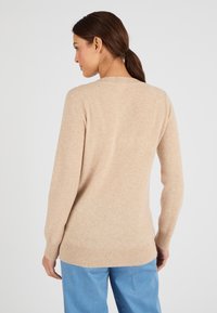 Maglione a maglia beige con maniche lunghe e orlo a coste. Caratterizzato da un collo rotondo e una texture morbida. Indossato con pantaloni azzurri chiari.