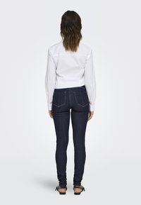 Witte longsleeve shirt, slim fit, gecombineerd met donkerblauwe skinny jeans met achterzakken en minimale stiksel. Sandalen zichtbaar bij de enkels.