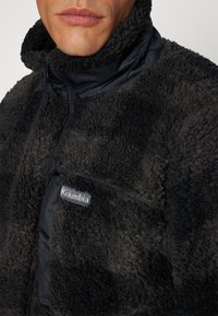 Giacca di fleece nera con superficie texturizzata, colletto alto e tasca con zip dotata di una patch con logo sulla parte anteriore. Materiale morbido e caldo.