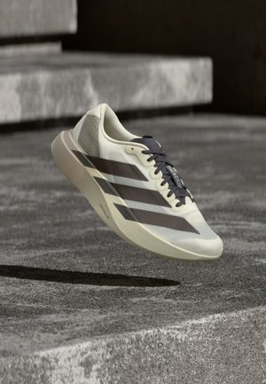 Chaussure de course Adidas Adizero beige clair et noire en lévitation au-dessus d'une surface en béton texturée avec des marches en pierre en arrière-plan.