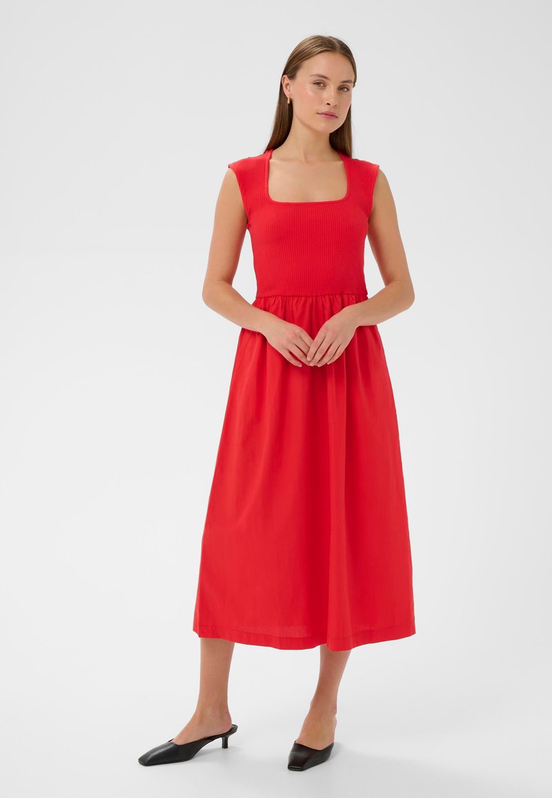 Punane, ilma varrukat maxi kleit ruudukujulise kaelusega ja kogutud vöökoht. Sile kangas, voolav seelik. Kandmisele on lisatud mustad kontsakingad.