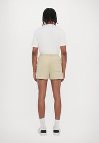 Beige Shorts mit einem verstellbaren Bund und einer einzelnen Gesäßtasche, kombiniert mit einem weißen T-Shirt. Der Stoff wirkt leicht und glatt.