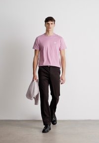 Lys rosa kortermet t-skjorte med en liten logo-lomme, kombinert med svarte bukser og svarte sko, holder en lys grå jakke i venstre hånd.