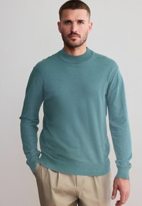 Teal crewneck sweater med lange ærmer, lavet af glat stof, med ribbet krave og kant. Båret med lyse bukser.