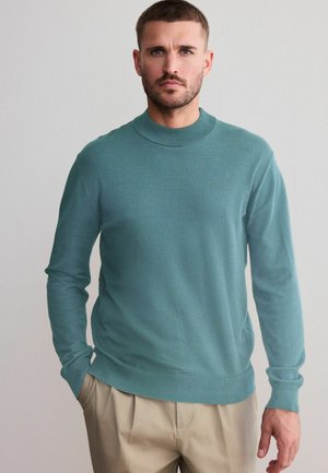 Teal crewneck sweater med lange ærmer, lavet af glat stof, med ribbet krave og kant. Båret med lyse bukser.
