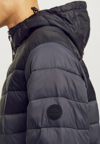Padded black jacket with a hood, featuring a smooth texture, horizontal quilted pattern, and a round logo patch on the sleeve.