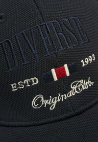 Granatowa czapka baseballowa z teksturowanej tkaniny, z dużym haftowanym logo "DIVERSE" oraz akcentującymi paskami w kolorach czerwonym, białym i niebieskim. Znajduje się także napis "ESTD 1993" oraz "Original Cloth".