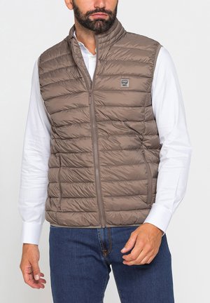 Uomo che indossa un gilet trapuntato marrone sopra una camicia bianca a maniche lunghe e jeans blu, in piedi contro uno sfondo chiaro e uniforme.