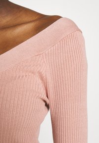 Růžový žebrovaný pletený top s designem bez ramínek. Má široký výstřih a texturovanou tkaninu, která zdůrazňuje hladký, jemný povrch.