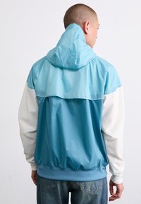 Viegls jacket ar zilu un balto krāsu bloku dizainu, ar kapuci un elastīgām aprocēm, izgatavots no gludas, spīdīgas materiāla.