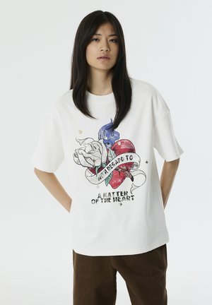 Witte t-shirt met een grafisch ontwerp van een hart, een roos en blauwe details, met de tekst "A Matter of the Heart." Korte mouwen en een ontspannen pasvorm.