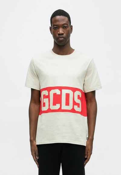 T-shirt beige a maniche corte con una banda rossa prominente con lettere bianche "GCSD" attraverso il petto. Materiale in cotone, vestibilità rilassata.
