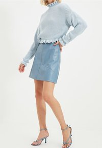 Kobieta ubrana w jasnoniebieski sweter z falbankami, pasującą niebieską skórzaną mini spódnicę i niebieskie sandały na obcasie, stojąca z ręką na biodrze.