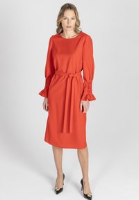 Czerwona sukienka z okrągłym dekoltem, długimi rękawami zakończonymi falbankami, dopasowaną talią z paskiem oraz długością do kolan.