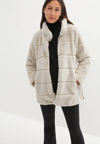 Giacca in pelliccia sintetica color beige chiaro, caratterizzata da un design trapuntato con strisce orizzontali, colletto, tasche laterali e una vestibilità rilassata.