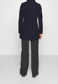 Cappotto lungo navy con silhouette aderente, caratterizzato da un colletto alto e maniche dritte, abbinato a pantaloni ampi scuri e calzature nere.