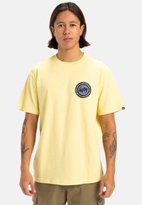 Camiseta de algodón amarilla con mangas cortas, que presenta un logo circular en azul marino y púrpura en el lado izquierdo del pecho. Corte relajado, diseño minimalista.