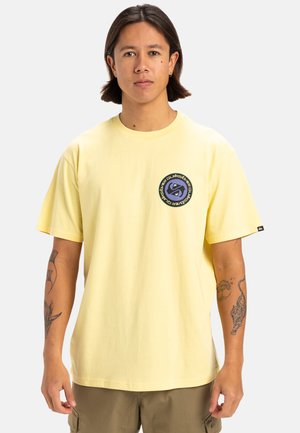 Camiseta de algodón amarilla con mangas cortas, que presenta un logo circular en azul marino y púrpura en el lado izquierdo del pecho. Corte relajado, diseño minimalista.
