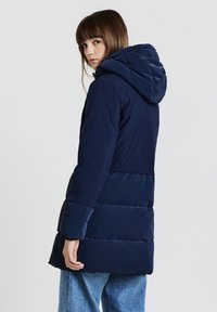 Cappotto imbottito blu navy con cappuccio, caratterizzato da pannelli testurizzati a contrasto e maniche lunghe. Il tessuto appare liscio e resistente alle intemperie.