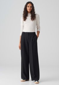 Cream long-sleeve top paired with high-waisted, wide-leg black trousers. The model wears black and white sneakers, displaying a relaxed fit.
