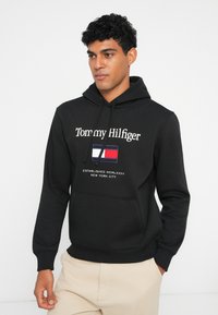 Schwarzer Kapuzenpullover aus Baumwolle mit einer Fronttasche. Mit weißem gesticktem Text und einem bunten Flaggenemblem auf der Brust.