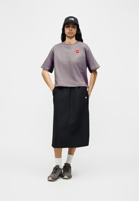 Γυναίκα που στέκεται με τα χέρια στις τσέπες, φορώντας μωβ κοντό μπλουζάκι, μαύρη μέση φούστα, λευκές κάλτσες, παπούτσια πεζοπορίας και μαύρο καπέλο.