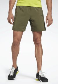 Olivově zelené sportovní šortky s hladkou strukturou, s bočními kapsami a černým logem na přední straně. Nosiči se černé tenisky.