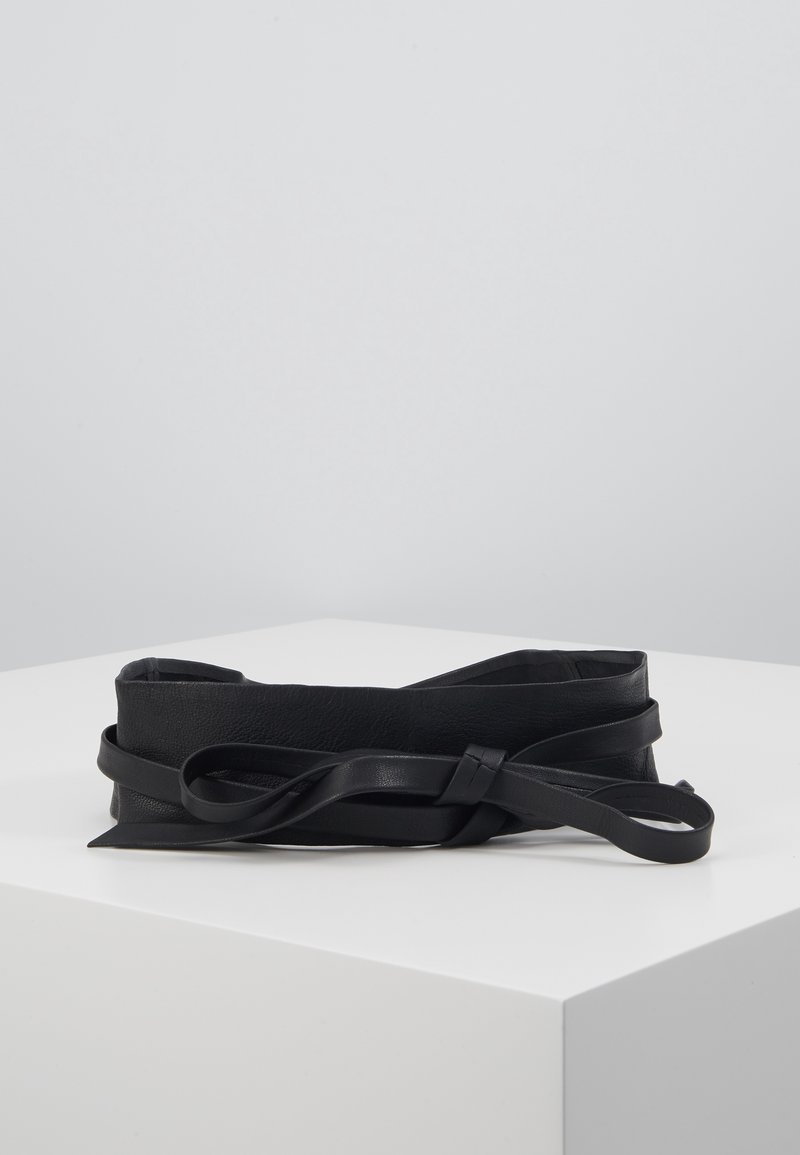 Ceinture de taille en cuir noir avec une longue sangle fine nouée en un nœud, exposée sur une surface blanche avec un fond gris uni.