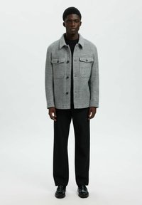 Joven de pie, vistiendo una chaqueta de lana gris abotonada sobre un suéter negro, pantalones anchos negros y zapatos de cuero negros sobre un fondo blanco.