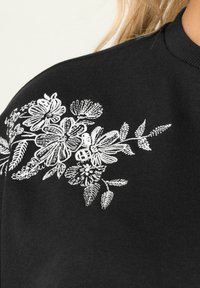 T-shirt nera con intricati ricami floreali bianchi sulla spalla, dal tocco morbido e design classico a girocollo.