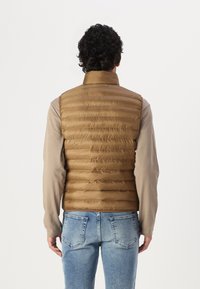 Vest puf, de culoare maro, cu guler înalt, având capse orizontale. Purtați peste o cămașă crem cu mânecă lungă; asortat cu blugi de culoare albastru deschis.