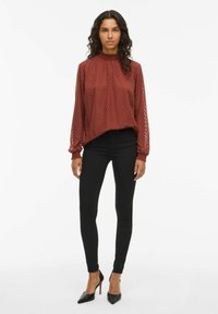 Bordeaux långärmad blus med hög hals, texturerat chevronmönster och elastiska ärmslut, kombinerad med svarta åtsittande byxor och svarta klackar.