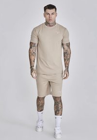 Šviesiai ruda trumpomis rankovėmis medvilninė marškinėliai ir atitinkami šortai, pagaminti iš minkštos medvilnės. Marškinėliuose yra subtilus logotipas, dėvimi su baltais sportbačiais ir kojinėmis.