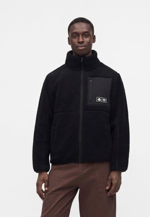 Junger Mann trägt eine schwarze Fleecejacke mit Reißverschluss und einer Reißverschlusstasche auf der Brust sowie braune Hose, steht vor einem weißen Hintergrund.