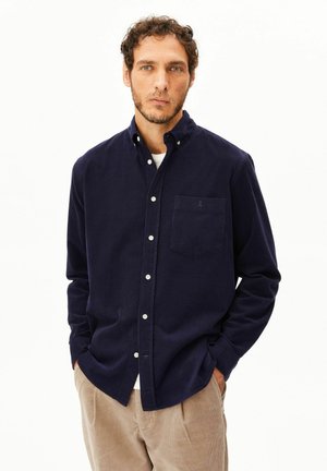 Mann mit kurzen lockigen Haaren trägt ein marineblaues Hemd mit Knöpfen über einem weißen Unterhemd und beigen Hosen, steht mit den Händen in den Taschen vor weißem Hintergrund.