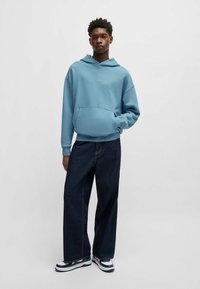 Felpa blu chiaro con tasca anteriore e cappuccio con cordino, abbinata a jeans larghi in denim scuro e sneakers blu e bianche.