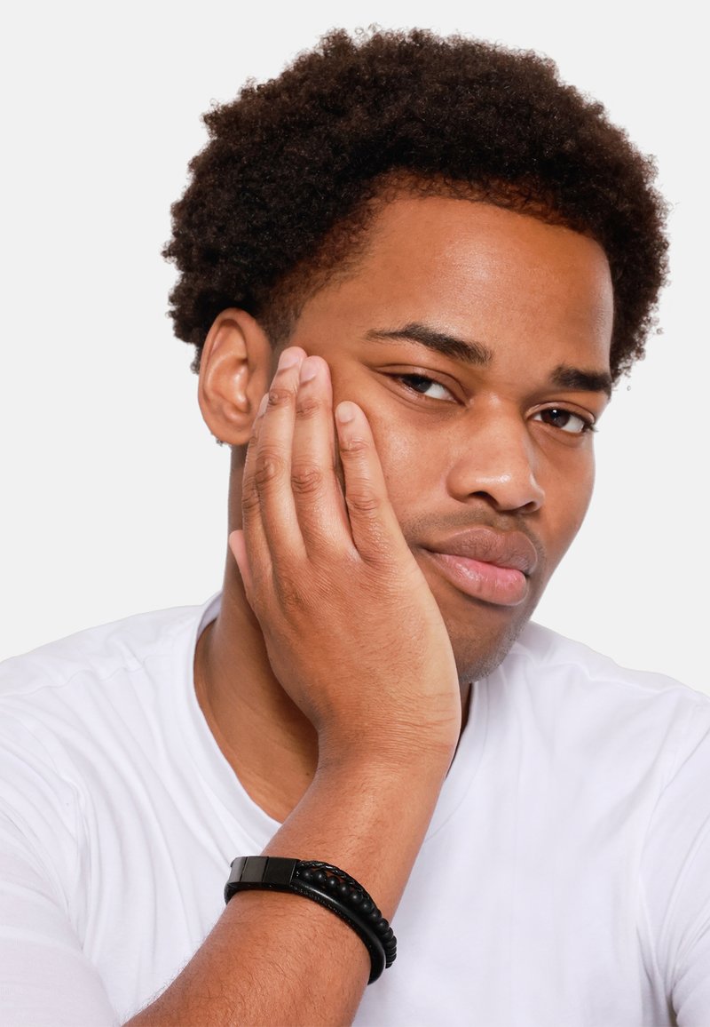 Giovane con capelli corti e ricci neri, indossa una t-shirt bianca. Riposa il mento sulla mano, mostrando un braccialetto di perline nere.