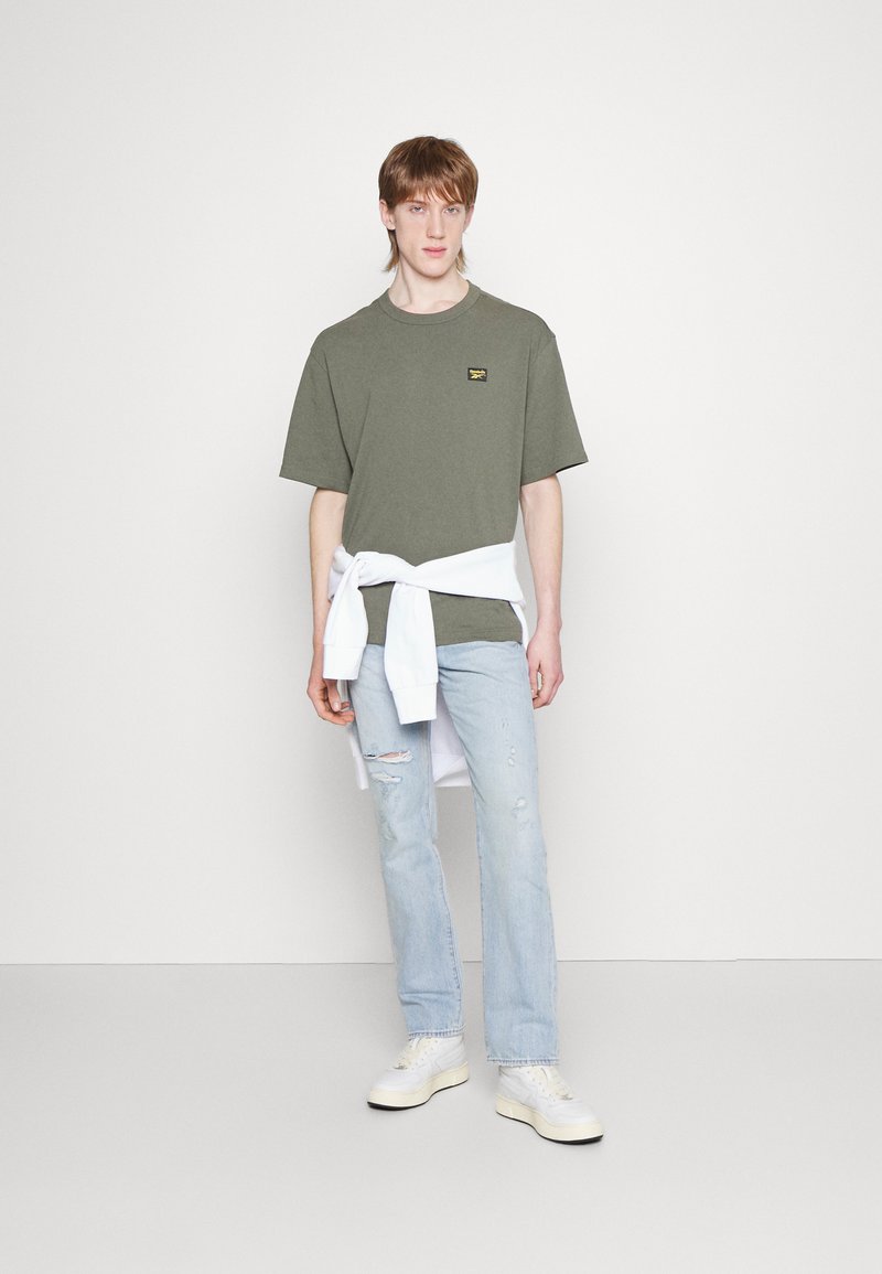 Olive green short-sleeve T-shirt with a small black and yellow logo, paired with light blue distressed jeans and white sneakers. White sweatshirt tied at the waist.