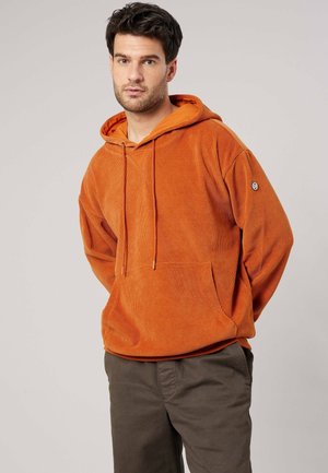 Man staat en draagt een oranje ribfluwelen hoodie met voorzak en een bruine broek tegen een effen lichte achtergrond.
